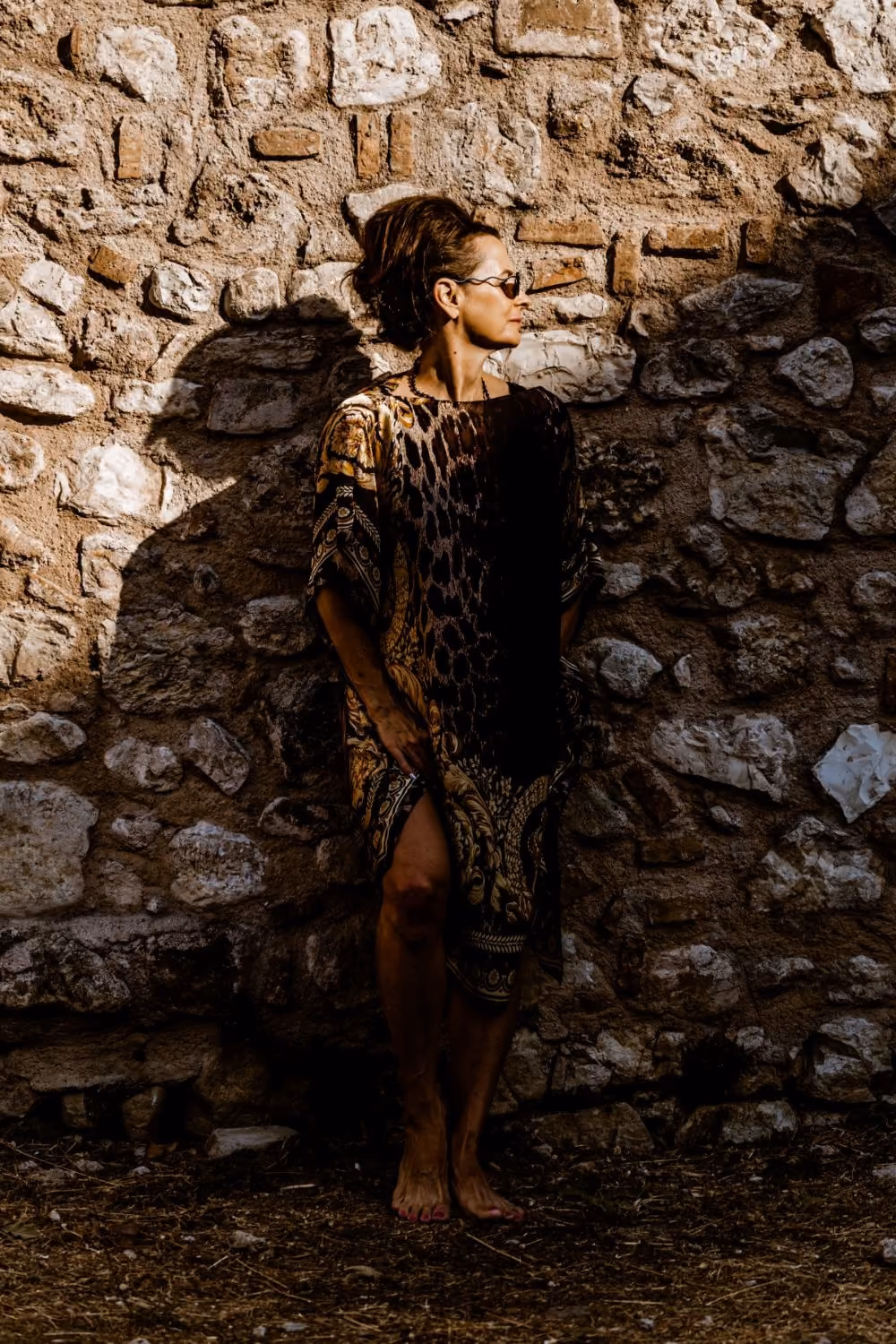Elle Wilson standing barefoot against an old stone wall in strong summer light, looking away to one side