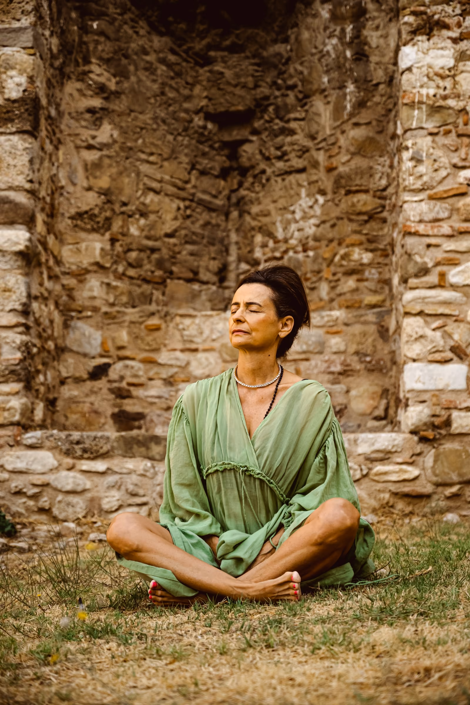 Elle Wilson seated cross-legged on the grass before an old stone wall, her eyes closed in meditation