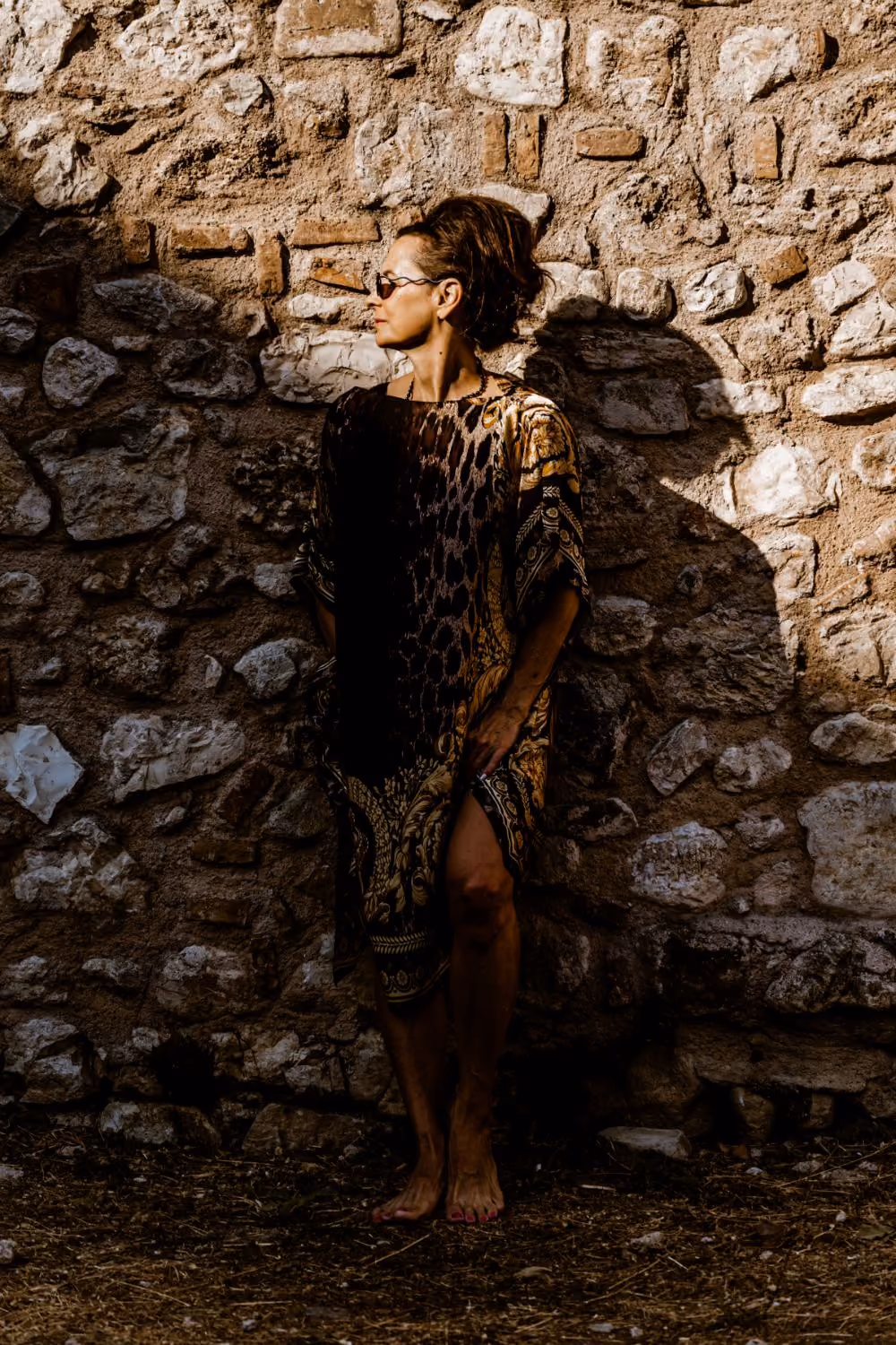 Elle Wilson standing barefoot against an old stone wall in a patterned kaftan, her face turned to one side